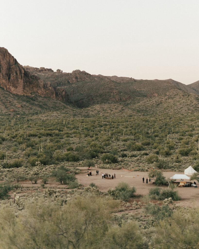 Aerial view of a desert landscape with a sparse gathering of people near a small white tent, surrounded by cacti and rocky hills in the background.