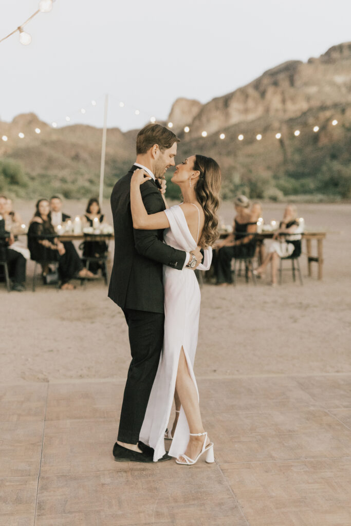 A couple in formal attire shares a dance outdoors on a wooden floor, with string lights above and seated guests in the background.