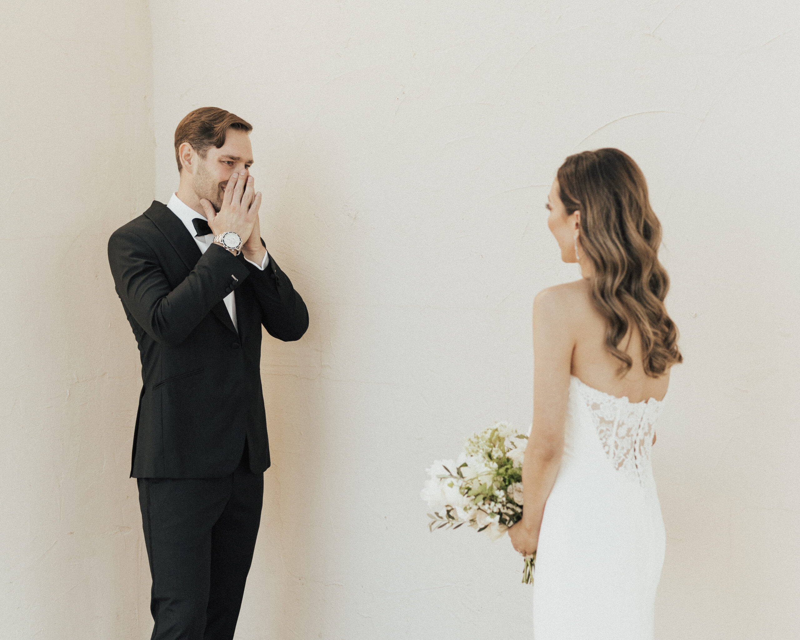 A groom in a black suit looks surprised and delighted as he sees the bride in a white dress holding a bouquet.