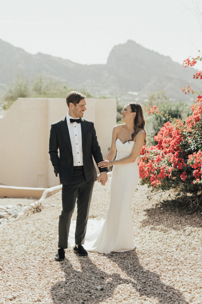 A bride and groom hold hands outdoors in a garden setting with bright red flowers and mountains in the background.