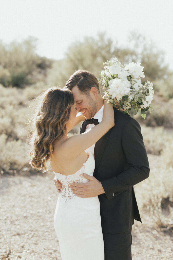 A couple in wedding attire embraces in a desert landscape. The bride holds a bouquet of white flowers.