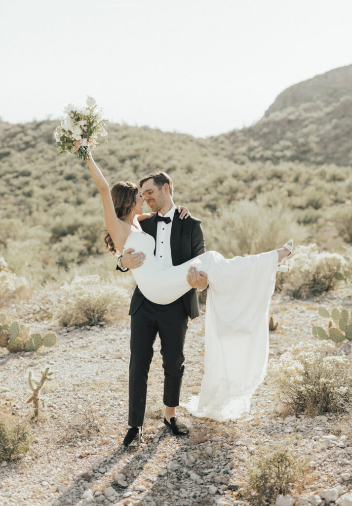 A groom in a tuxedo lifts the bride in a white dress, holding a bouquet, in a desert landscape with cacti and hills in the background.