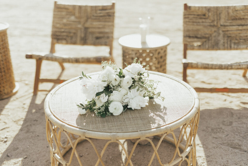 A bouquet of white flowers on a round wicker table. In the background, two wicker chairs and a small side table on a sandy surface.
