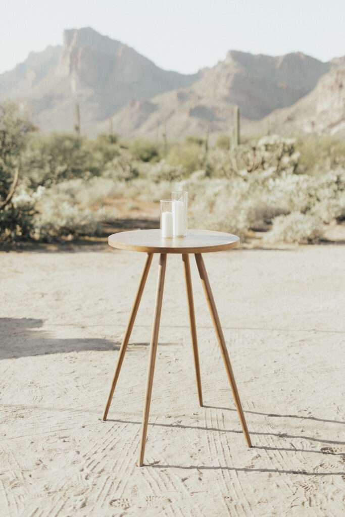 A wooden tripod table with a glass of milk stands on a sandy ground with cactus and mountains in the background.