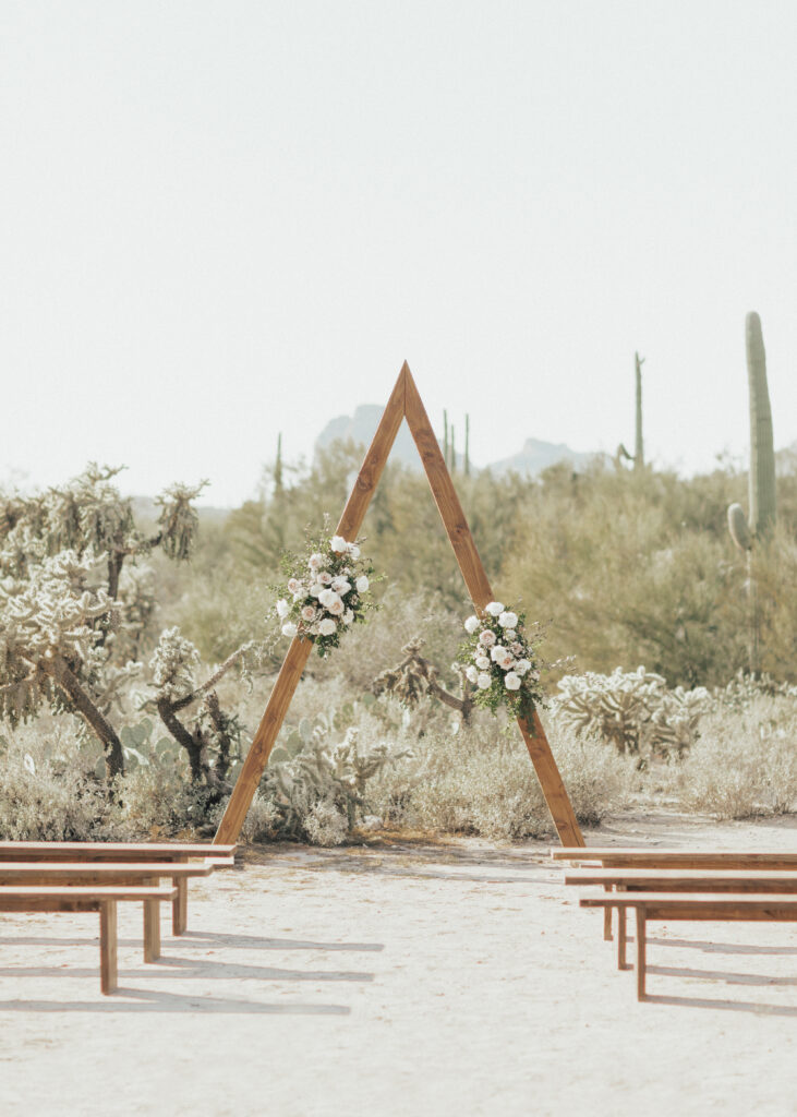 Wooden triangular arch with floral decorations set in a desert landscape with cacti, framed by wooden benches, under a clear sky.