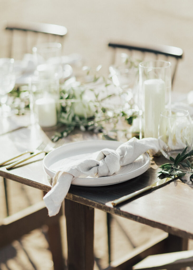 A wooden table set for dining with a white plate, rolled napkin, and greenery. Candles and glassware are arranged around it.