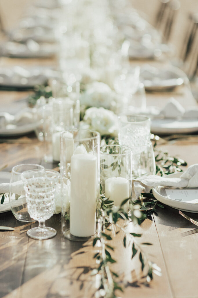 A long wooden table is decorated with white candles in glass holders, white flowers, and greenery. Plates and glassware are neatly arranged beside folded napkins.