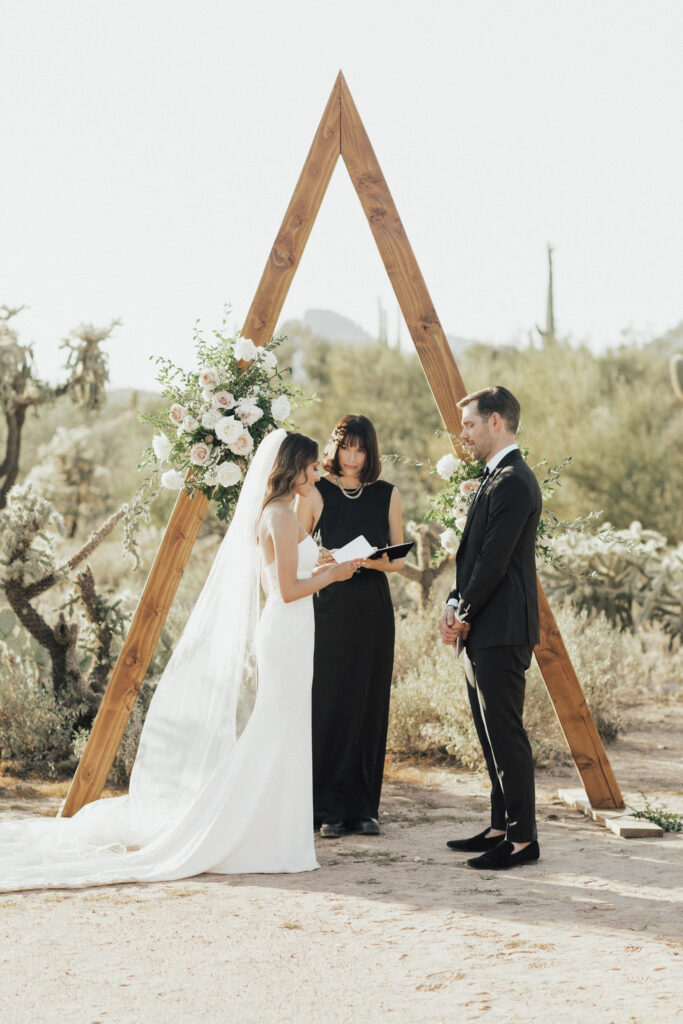 A bride and groom stand facing each other in front of a triangular wooden arch adorned with flowers. A woman officiates the ceremony outdoors in a desert setting.