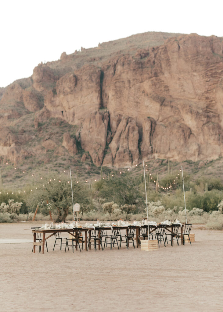 Outdoor dining setup on a sandy area, with long wooden tables and chairs. String lights are hung above, set against a backdrop of rocky mountains.