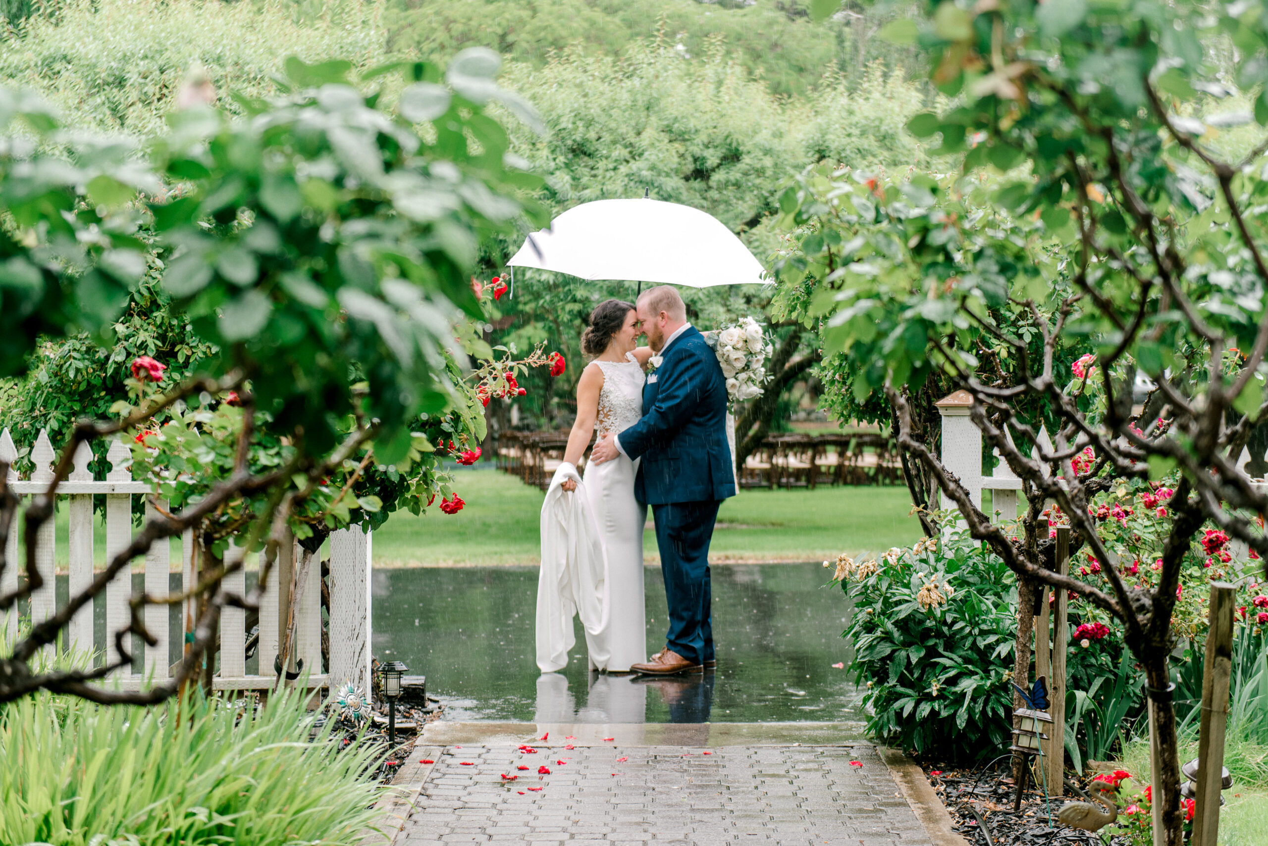 A couple dressed formally stands close together under a white umbrella on a rainy garden path, surrounded by green foliage and red flowers.