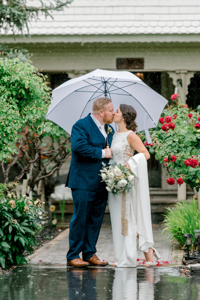 A bride and groom stand under a white umbrella, kissing in a garden with red roses, on a rainy day outside a white house.