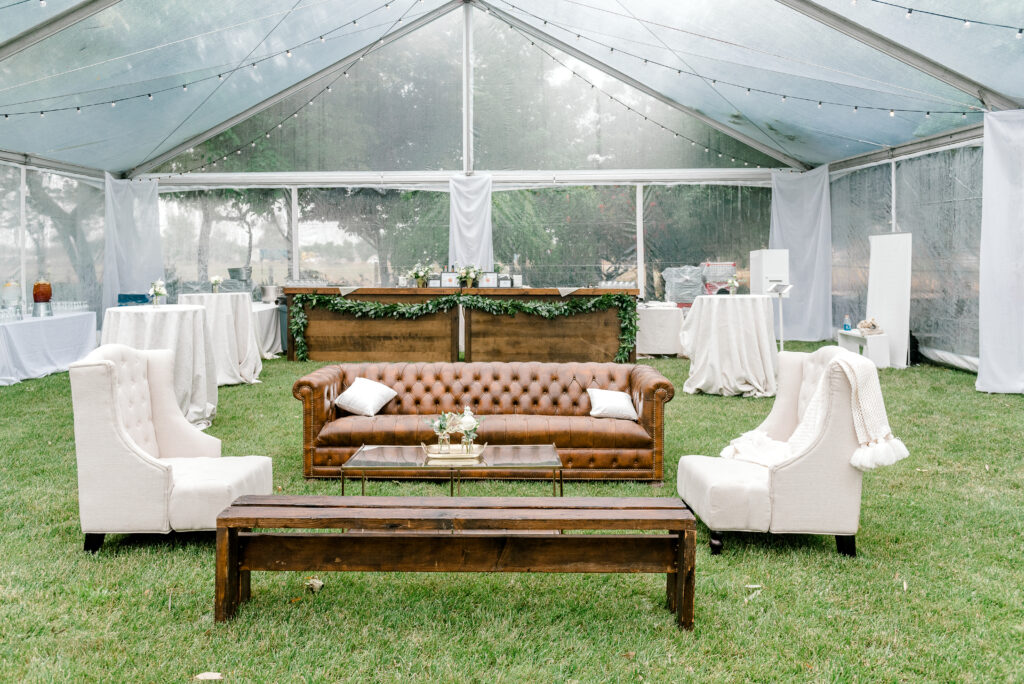 A clear tent set up outdoors with a brown leather sofa, two white armchairs, a wooden bench, and covered tables arranged on green grass.
