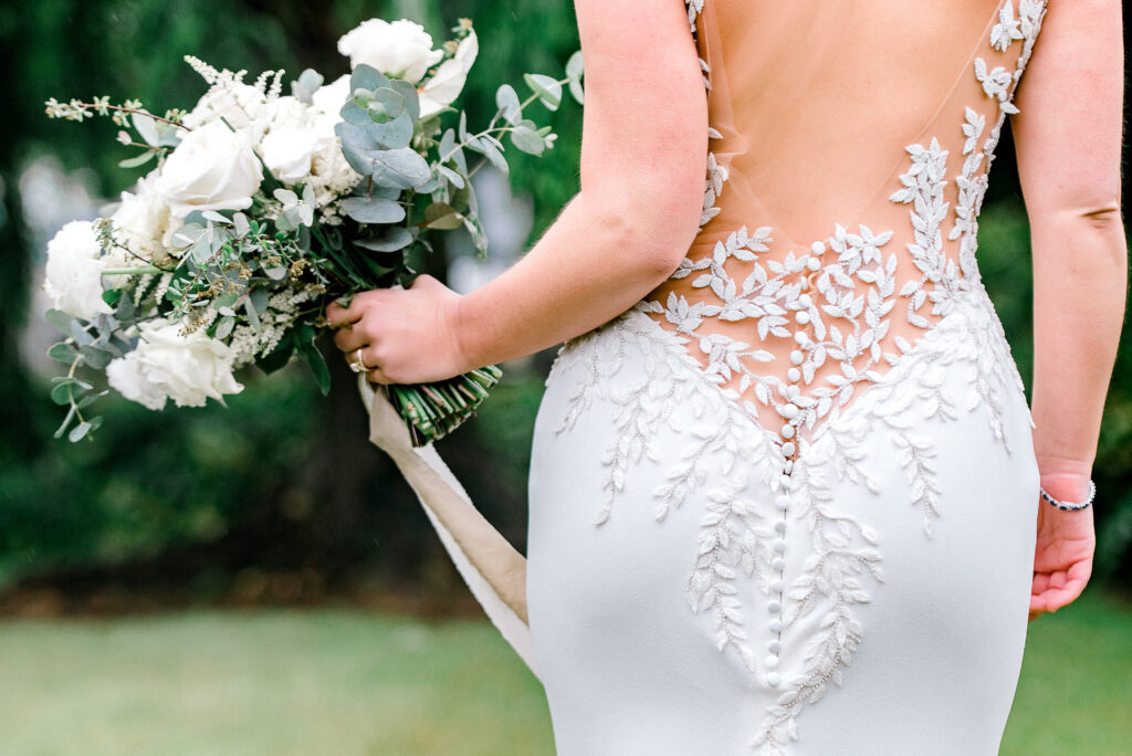 A person in a white lace wedding dress holds a bouquet of white flowers and greenery outdoors.