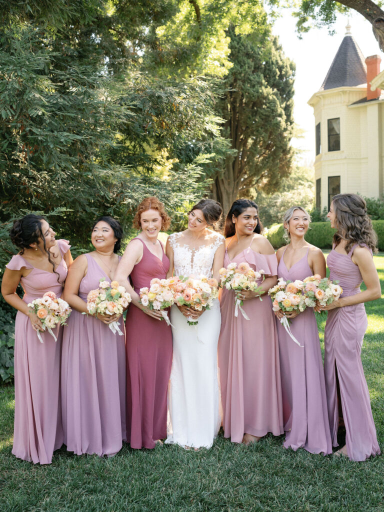 A bride in a white dress stands outdoors with six bridesmaids in pink dresses, all holding floral bouquets, in front of a tree and a Victorian-style house.
