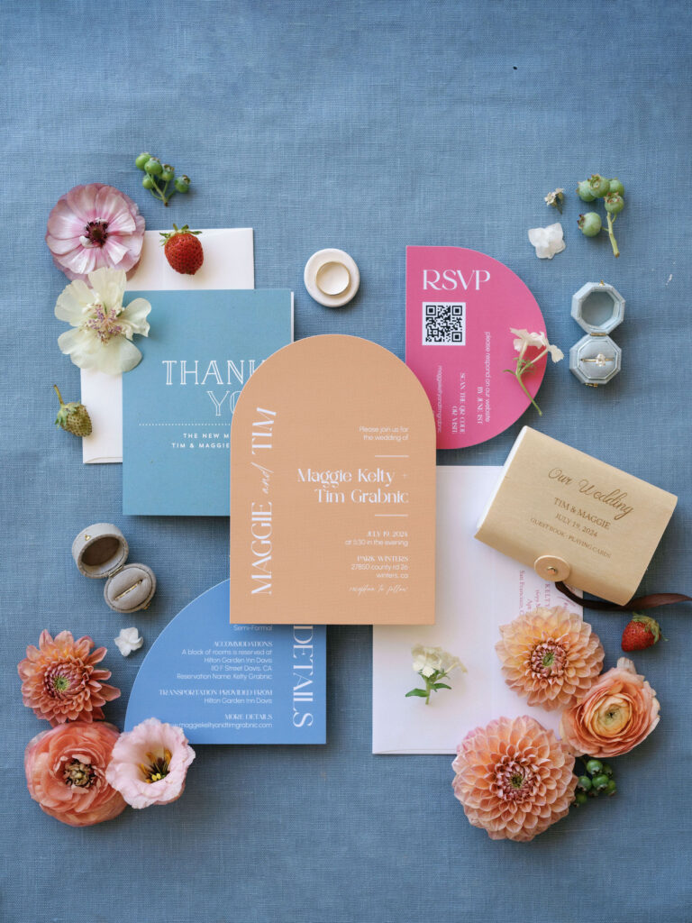 A flat lay of wedding invitations, thank you cards, and RSVP cards in various pastel colors, arranged with flowers, strawberries, and macarons on a blue surface.