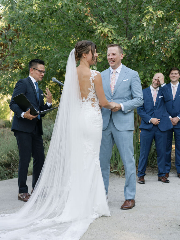 A couple stands facing each other during an outdoor wedding ceremony. The bride is in a white gown, and the groom is in a light blue suit. A person officiates and groomsmen are in the background.