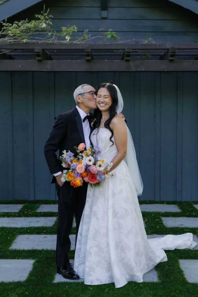 An older man in a suit kisses a smiling bride holding a colorful bouquet, standing outside in front of a dark wooden structure.