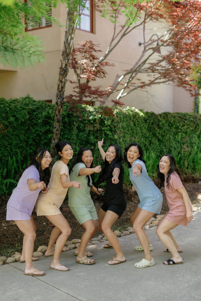 Six young women stand outdoors on a sidewalk, wearing casual matching outfits in different colors, smiling and pointing towards the camera. Shrubs and a building are in the background.