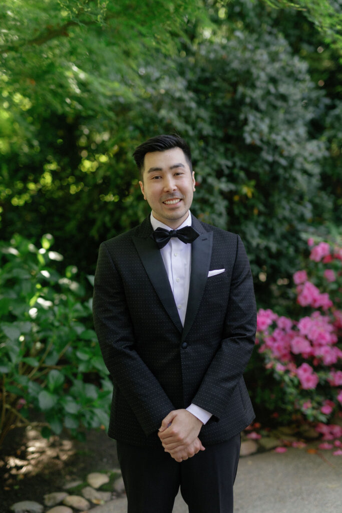 A man in a black tuxedo stands outdoors, surrounded by greenery and pink flowers, smiling at the camera.