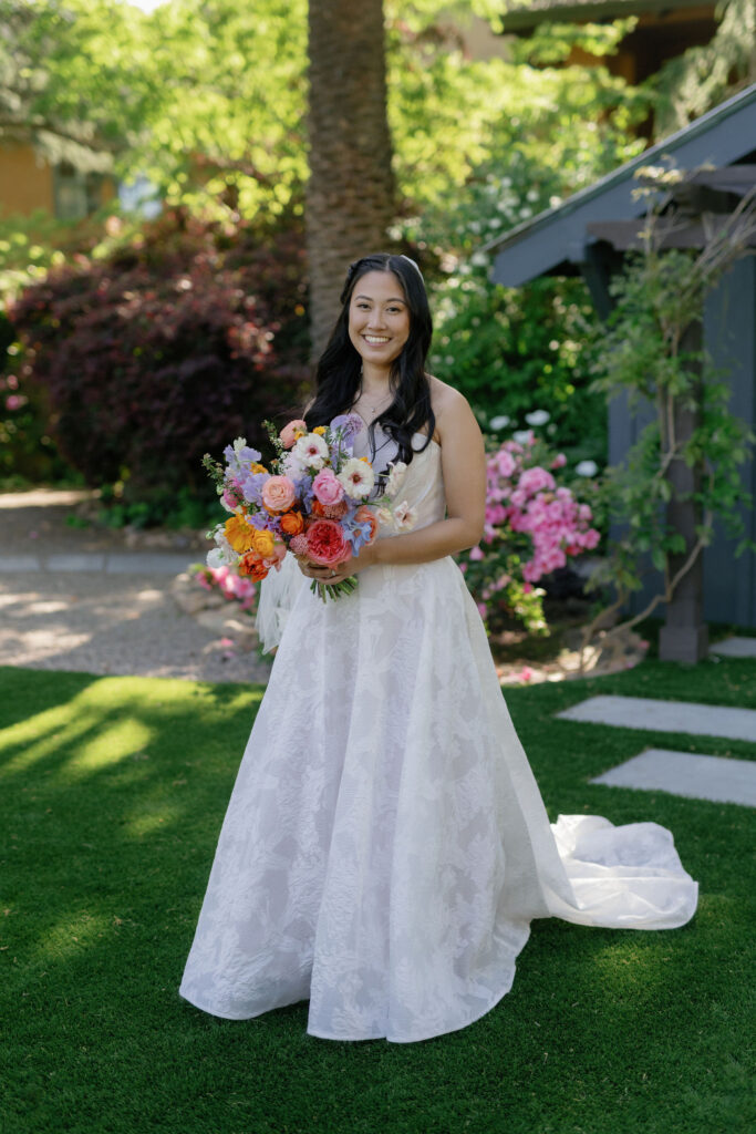 A woman in a white wedding dress stands on grass holding a colorful bouquet of flowers, with trees and blooming plants in the background.