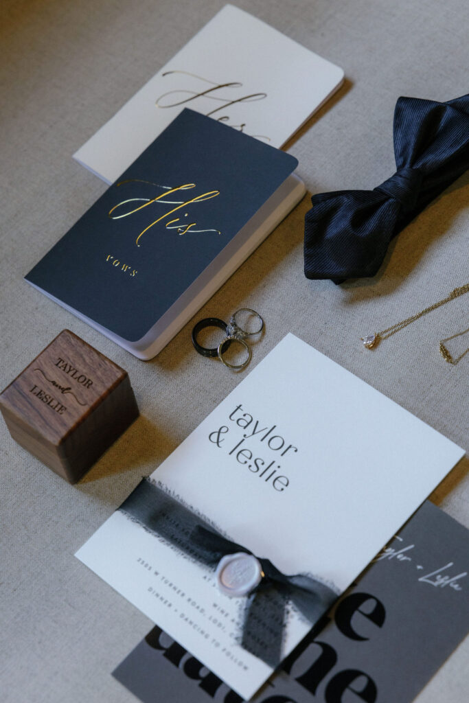 Wedding invitation suite with vow books, rings, a bow tie, a necklace, and a wooden ring box labeled "Taylor & Leslie" arranged on a gray fabric surface.