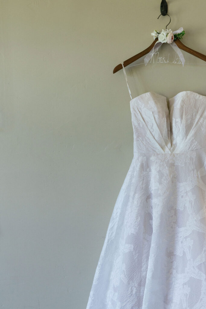 A white strapless wedding dress with a floral pattern hangs on a wooden hanger against a plain light-colored wall.