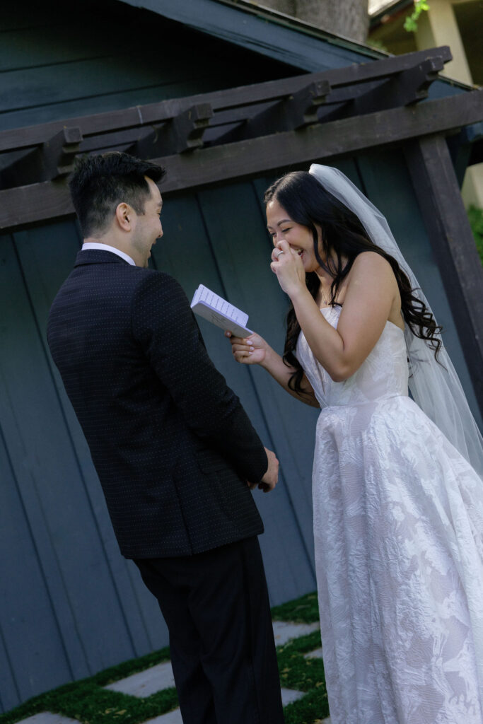 A bride in a white dress and veil reads from a piece of paper, smiling and covering her mouth, while facing a groom in a dark suit outdoors.