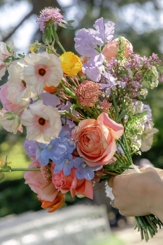 A hand holding a bouquet of assorted colorful flowers, including roses, blue, yellow, and pink blooms, with greenery visible in the background.