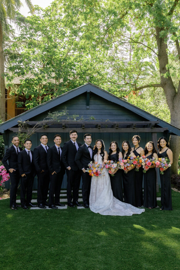 A bride and groom stand with their wedding party, all dressed in black formal attire, in front of a small blue building with trees in the background.