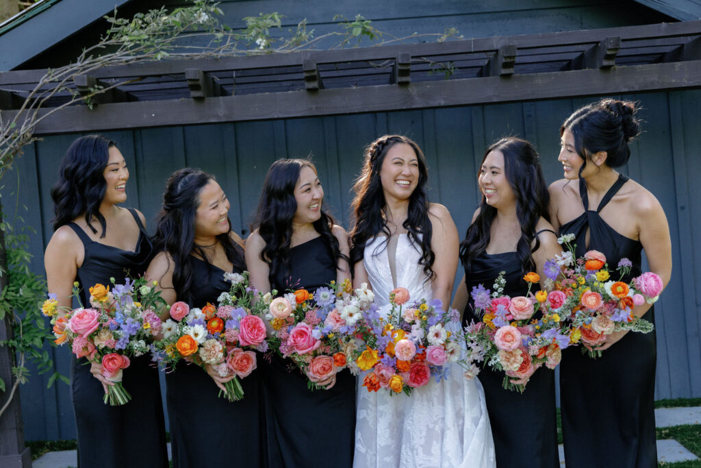 Six women stand together outdoors, one in a white dress and five in black dresses, all holding colorful flower bouquets and smiling.