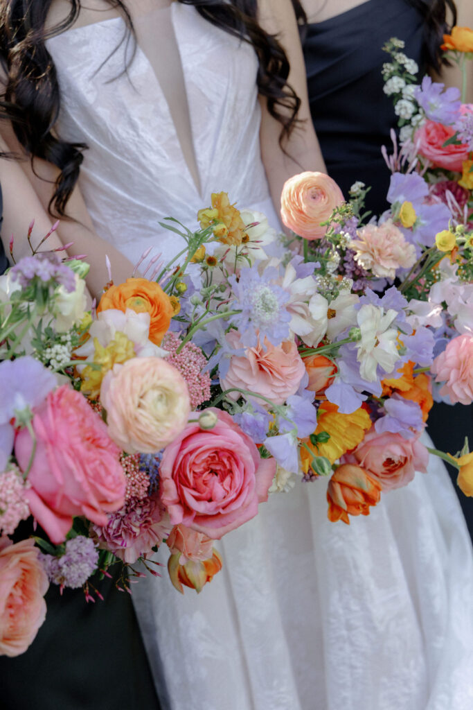 A bride in a white dress holds a colorful bouquet of assorted flowers, including roses, ranunculus, and delphiniums, next to others in dark dresses.
