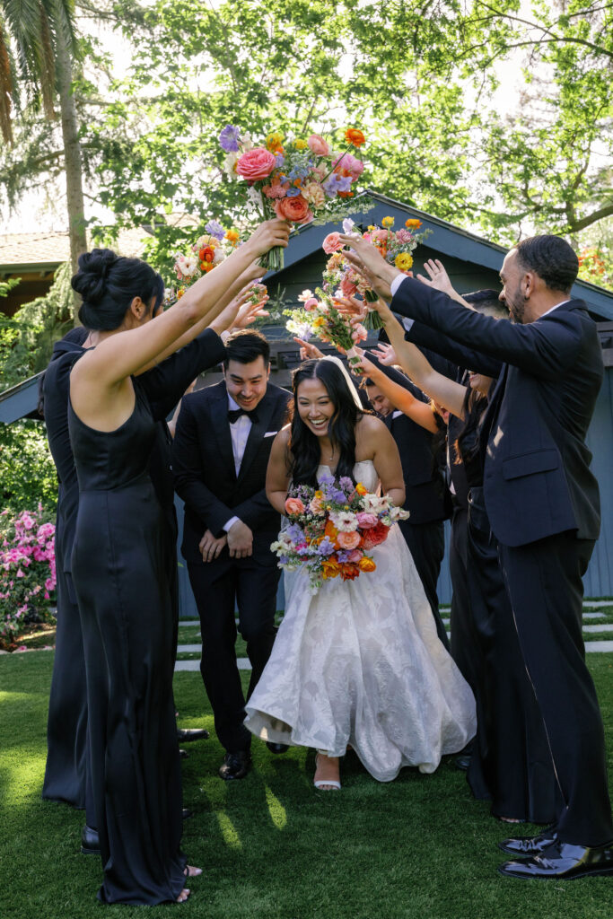 A bride and groom walk under an arch formed by attendants holding colorful flower bouquets; all are smiling outdoors in formal attire.