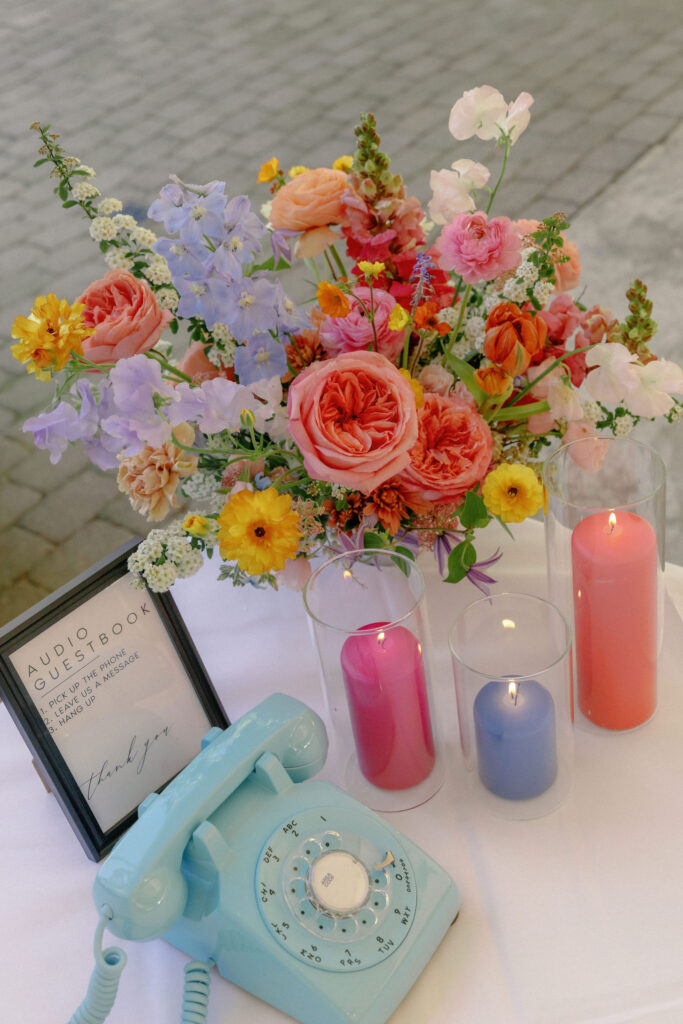 A pastel blue rotary phone, a framed audio guestbook sign, colorful flowers in a vase, and three lit candles sit on a white table.