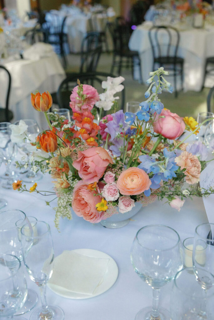 A floral centerpiece with pink, orange, and blue flowers sits on a white-clothed table surrounded by empty wine glasses at an event.