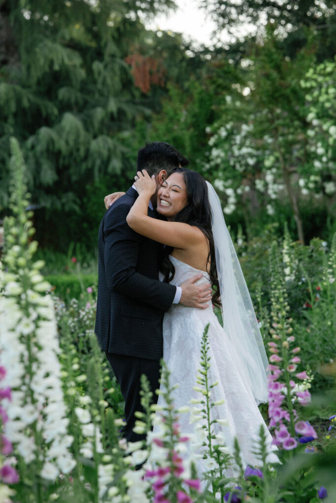 A bride in a white dress and veil hugs a man in a dark suit outdoors, surrounded by tall flowers and greenery.