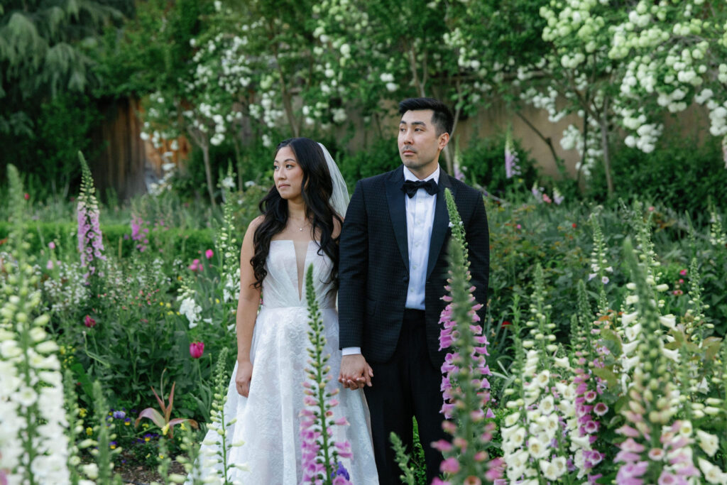 A bride and groom stand holding hands in a lush garden surrounded by tall flowering plants, both looking away from the camera.
