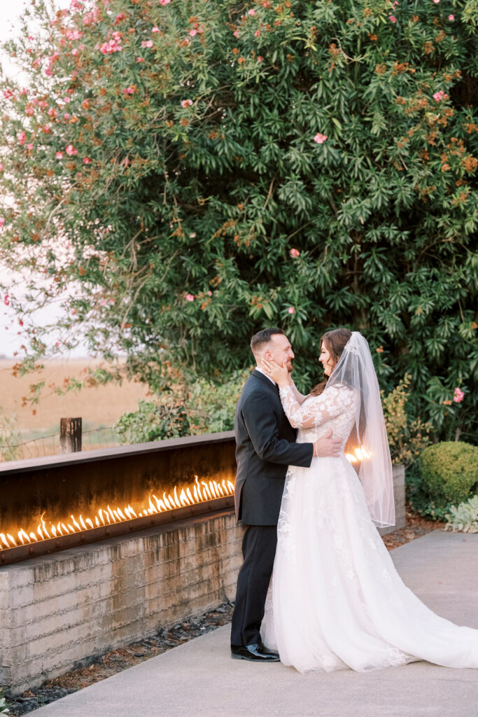 A bride and groom stand facing each other outside by a long fire feature, with greenery and flowers in the background.