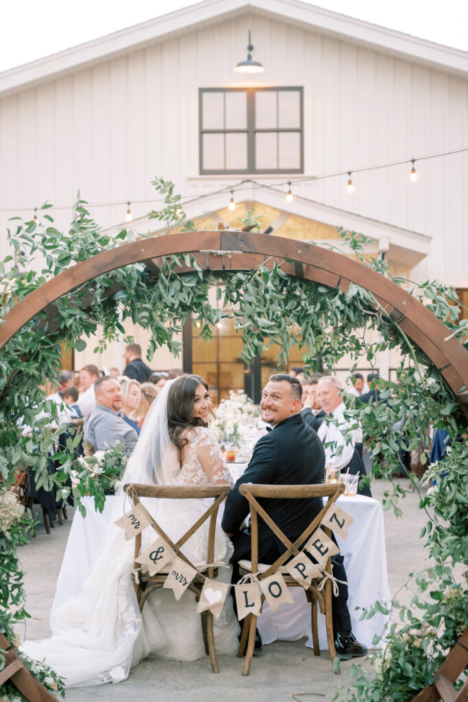 A bride and groom sit at a decorated outdoor table during their wedding reception, facing the camera, with a sign reading "Mr. & Mrs. Lopez" on their chairs.