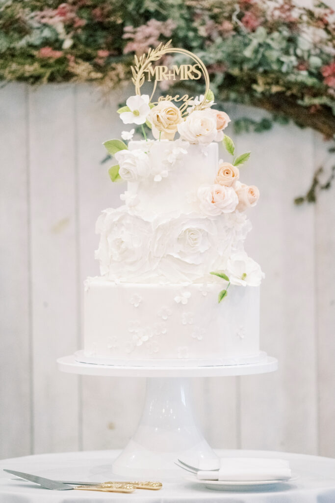 A white, two-tier wedding cake decorated with white and pale peach flowers, topped with a gold "Mr & Mrs" sign, displayed on a white cake stand.