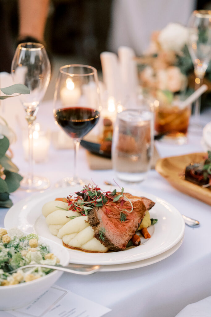 A plate of sliced beef and mashed potatoes is served on a white tablecloth with a glass of red wine, water, and a salad nearby.