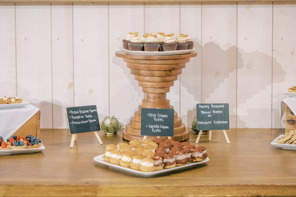 A dessert table displays cupcakes on a wooden stand, assorted pastries on trays, and small chalkboard signs listing dessert options against a white wooden backdrop.
