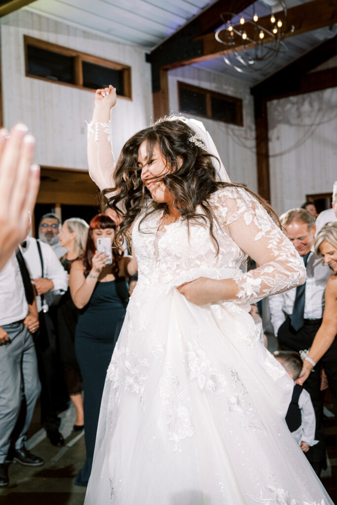 A bride in a white lace wedding dress dances joyfully among guests at a wedding reception, holding up her dress with one hand.