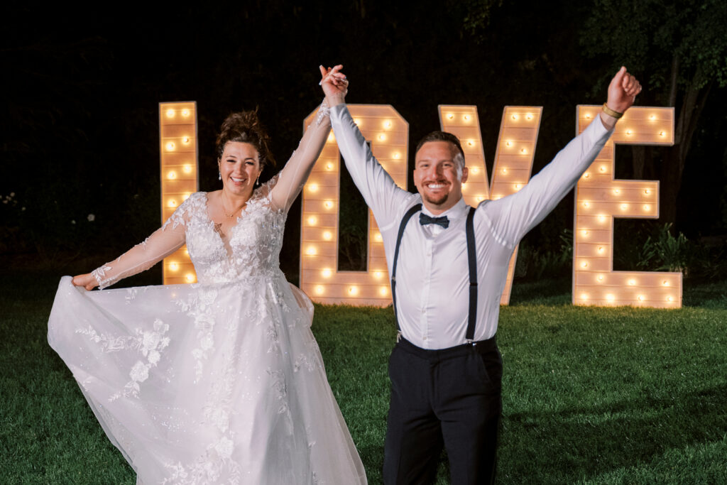 A bride and groom stand smiling with raised arms in front of large, illuminated "LOVE" letters on a grassy area at night.