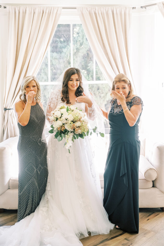 A bride in a white gown stands between two women in formal dresses, all wiping away tears, in front of a window with cream curtains. The bride holds a bouquet of white and pale flowers.