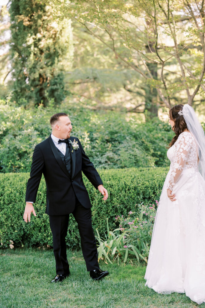 A groom in a black suit and a bride in a white gown with lace sleeves stand facing each other outdoors in a garden setting.