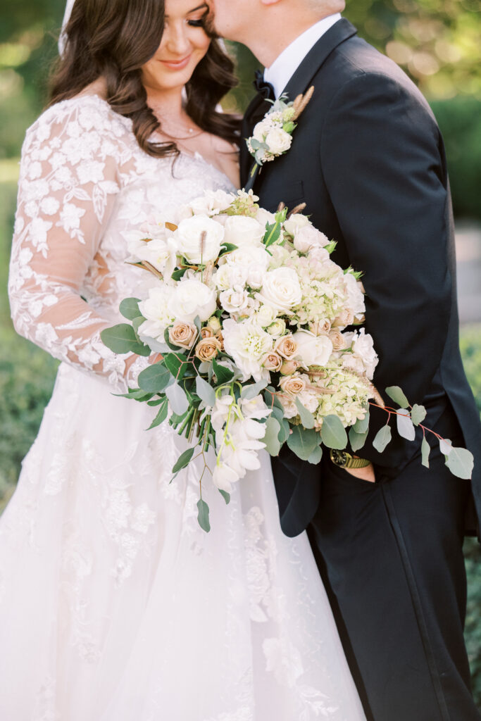 A bride in a lace wedding dress holds a large bouquet of white and pale flowers while standing beside a groom in a black suit.
