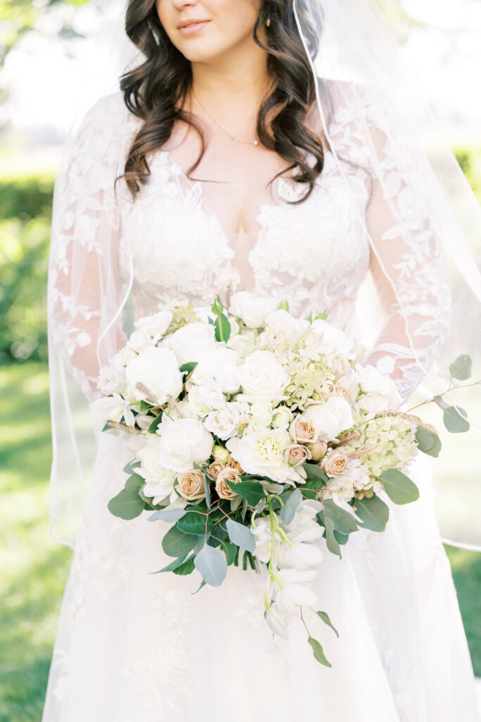 A bride in a white lace wedding dress holds a bouquet of white and pale pink flowers with greenery.