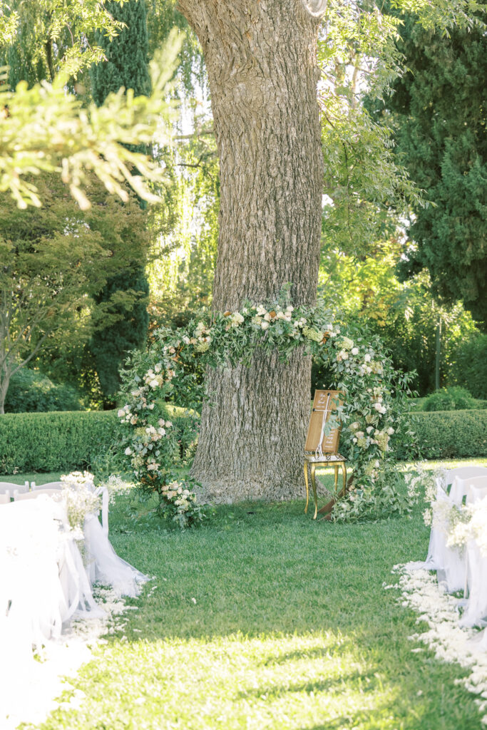A decorative floral arch and a single chair are set up outdoors in front of a large tree, with rows of white chairs on either side of an aisle covered in white petals.