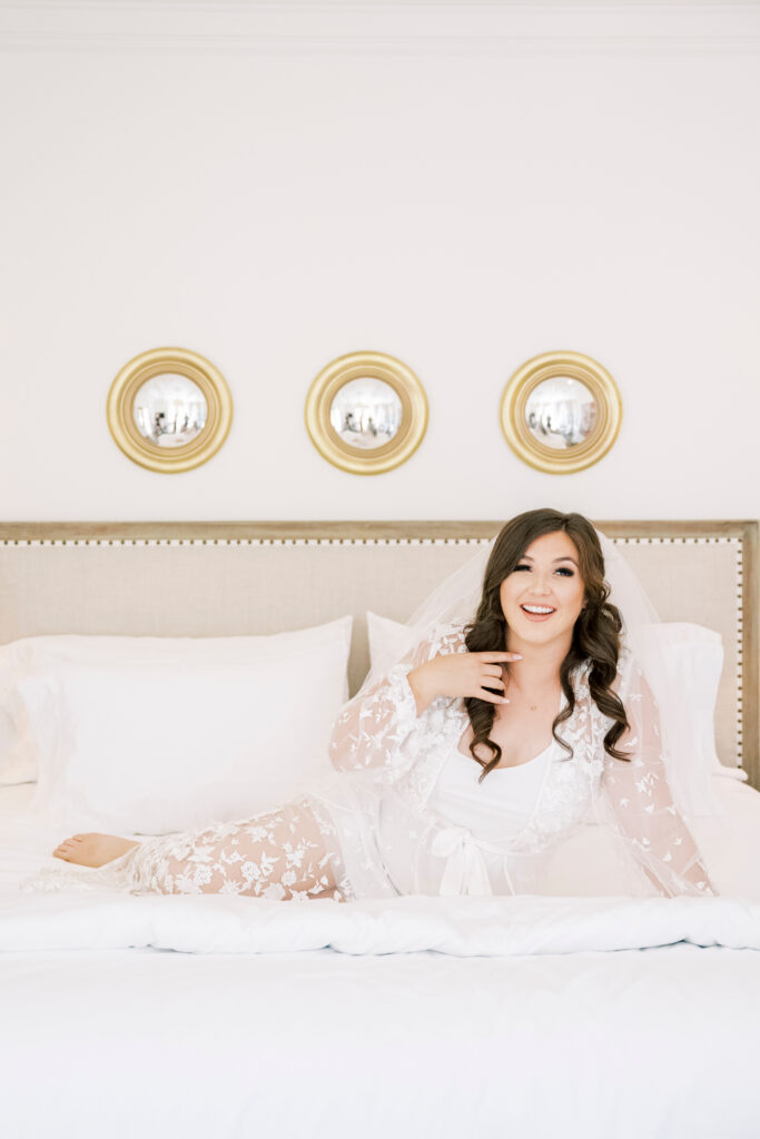 A woman in a white lace robe sits on a neatly made bed, smiling, with three round mirrors on the wall behind her.