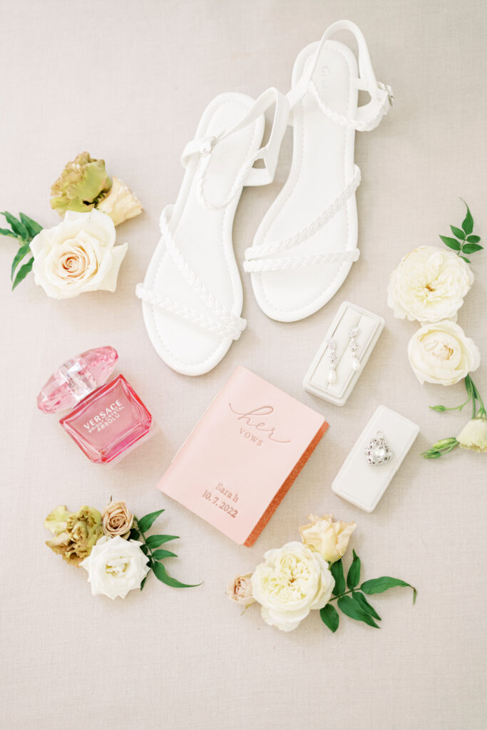 Flat lay of white sandals, floral arrangements, Versace perfume, a pink vow booklet, earrings, and rings on a light background.
